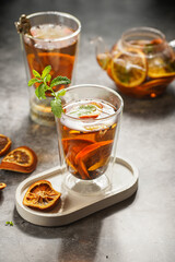 A transparent tea glass filled with black tea, oranges and mint on a grey concrete surface