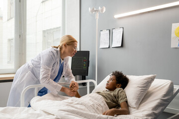 Obraz premium Female doctor in white coat caring about little boy who lying on bed after operation in hospital ward