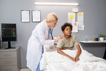 Obraz premium Doctor examining the lungs of little boy with stethoscope during medical exam in hospital ward