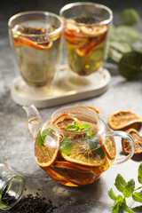 A transparent tea pot filled with black tea, oranges and mint on a grey surface