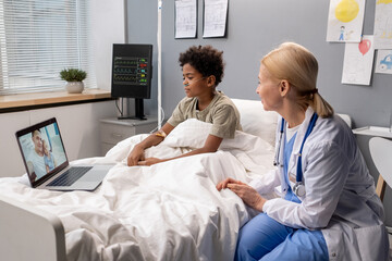 Sick little child sitting in bed together with his doctor and talking online with his family on computer at hospital ward