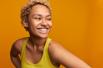 Close up of mixed-race cheerful gorgeous woman with natural beauty wearing short curly hair dyed in blonde, laughing showing white healthy teeth looking aside isolated on orange, dressed in yellow