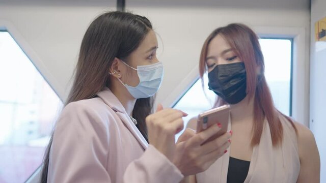 Two asian female closed colleague standing cheerfully talking while using a pubic train to office, attractive red dyed hair fashionista shows smartphone to friend, people working on covid-19 pandemic 