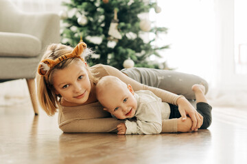 A charming girl and her younger brother play together on the floor next to the Christmas tree on Christmas or New Year's Eve