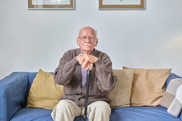Portrait of senior man in eyeglasses sitting on sofa with crutch and looking at camera during his...