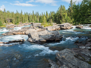 Beautiful northern landscape with rapids and cascades of wild river Kamajokk, boulders and spruce tree forest and hills in Kvikkjokk village in Swedish Lapland. Summer sunny day, blue sky white clouds