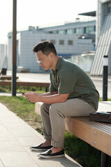 Fototapeta premium Young Asian businessman scrolling in smartphone while sitting on bench in urban environment on summer day