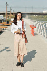 Young Chinese businesxswoman with drink standing in front of camera against riverside and large park
