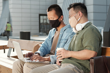 Two young Chinese businessmen in casualwear and protective masks preparing presentation while one of them typing