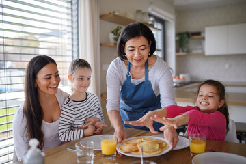 Happy small girls with mother and grandmother eating pancakes indoors at home, having lunch.