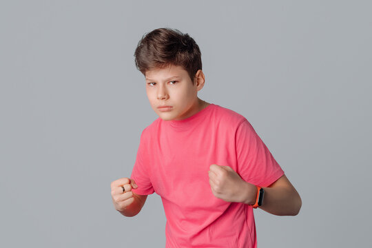 Portrait of annoyed angry teenager boy tight fists, feeling furious wearing casual pink t shirt standing over gray isolated background