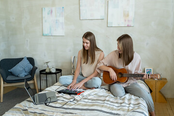 Two happy teenage girls recording music at home while one of them playing guitar and the other...