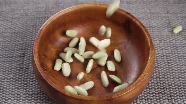Uncooked green beans fall into a wooden bowl on a gray rustic jute mat close up. Raw legumes. Traditional Spanish mediterranean cuisine