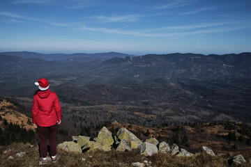 Woman in red Santa hat celebrates Christmas and New Year on top of mountain without snow in warm country. Stand with back turned and enjoy scenery and wildlife views. Tourist travels in red jacket.