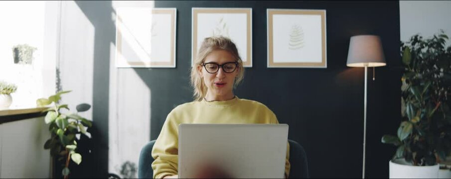 Zoom in shot of young woman in glasses sitting in armchair at home, waving and chatting on online video call on laptop