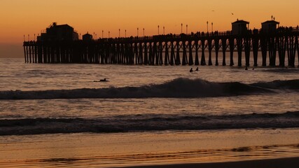Obraz premium Wooden pier silhouette at sunset, California USA, Oceanside. Waterfront surfing resort, pacific ocean tropical beach. Summertime sea coastline vacations atmosphere. Surfers waiting for wave in water.