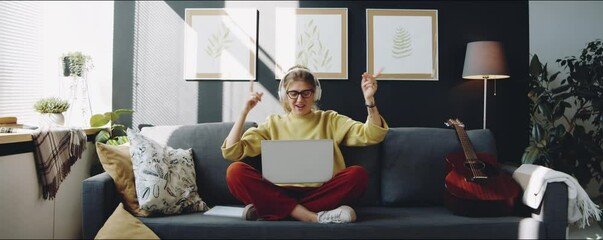 Zoom out shot of young woman in headphones sitting with legs crossed on sofa in modern living room, listening to music on laptop and dancing - Powered by Adobe
