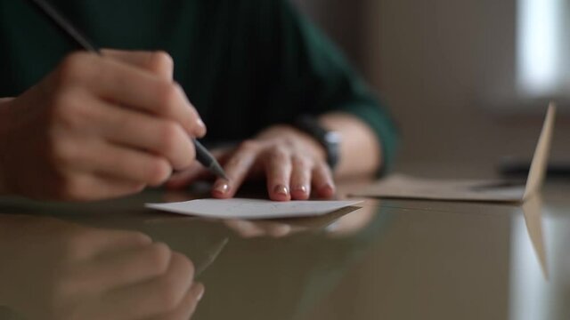Close-up hands of unrecognizable young woman writing handwritten wishes on Christmas card sitting at table at home, selective focus. Happy lady writes letter to boyfriend in love on Valentines Day.