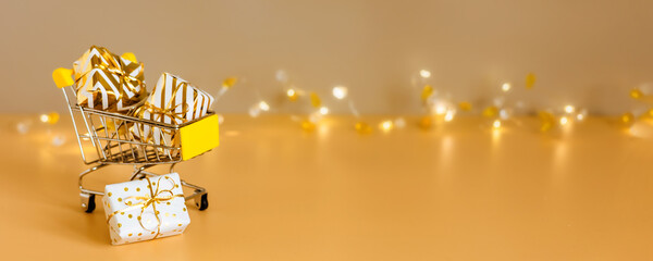 Online christmas shopping and sale. Shopping cart with gift boxes on golden background with lights. Supermarket trolley full presents. Banner with copy space