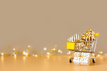 Online christmas shopping and sale. Shopping cart with gift boxes on golden background with lights. Supermarket trolley full presents. Copy space