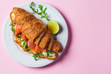 Croissant sandwich with cream cheese, salmon and arugula on white plate, pink background. Healthy...