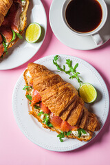 Croissant sandwich with cream cheese, salmon and arugula on white plate, pink background. Healthy breakfast concept.