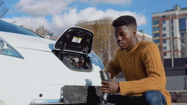 Dark-skinned male driver connects electric car to power system to charge car battery