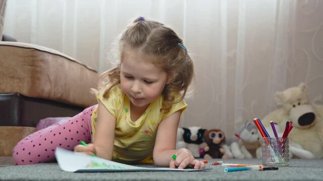 A little girl draws with felt-tip pens on paper, lying on the floor at home. Art, education, childhood, concept. High quality 4k footage