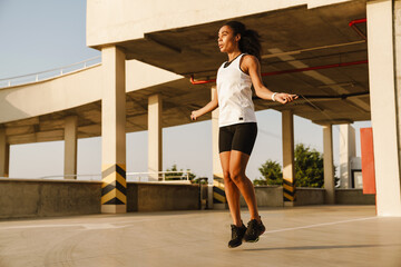 Black sportswoman working out with jumping rope on parking