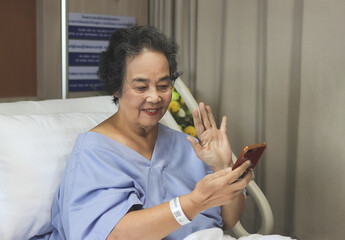 cheerful  Asian senior  female patient  sitting  in hospital bed,  smiling and looking at mobile phone, video calling  and waving to her family. Elderly health concept.