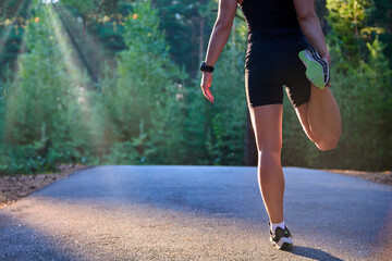 Athlete-runner, stretching her legs and feet and preparing to run on an asphalt road in the forest. Active, running training in the rays of sunlight and the concept of a healthy lifestyle in sports.