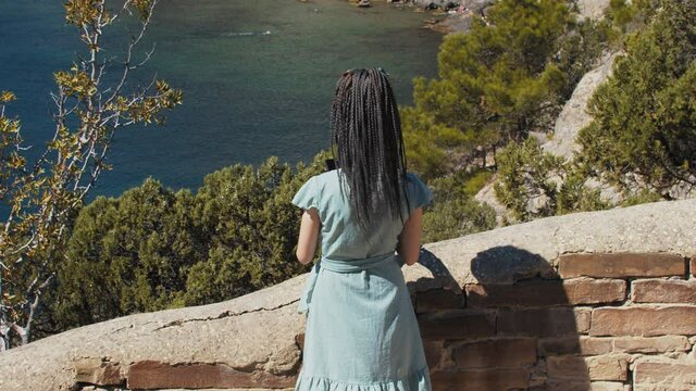 A young woman with dreadlocks walking down to the brick wall and taking a picture on her phone of the sea bay