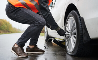 Side cropped view of man in black gloves and orange vest replacing tire after puncture on road,...
