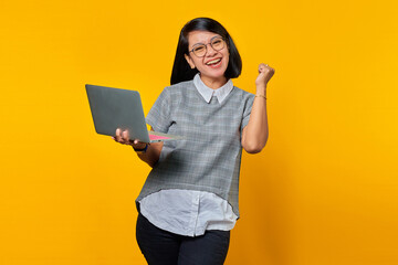 Portrait of excited Asian woman holding laptop over yellow background