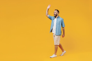 Full body young smiling happy cheerful caucasian man 20s wearing blue shirt white t-shirt walk going stroll waving hand isolated on plain yellow background studio portrait. People lifestyle concept