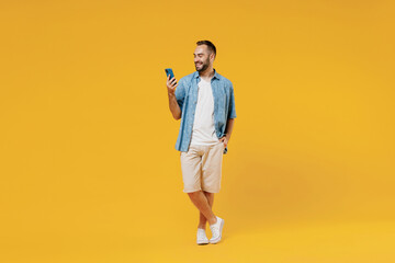 Fototapeta na wymiar Full body young smiling happy cool caucasian man 20s wearing blue shirt white t-shirt hold in hand use mobile cell phone isolated on plain yellow background studio portrait. People lifestyle concept.