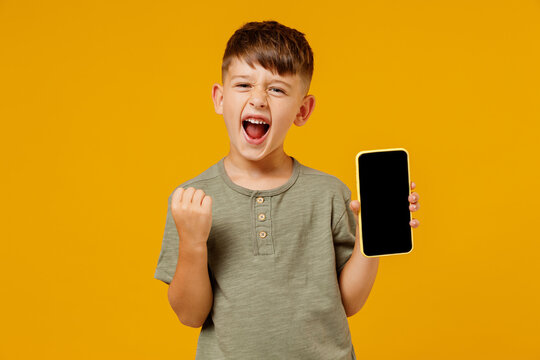 Little happy fun boy 6-7 years old in green t-shirt hold use mobile cell phone with blank screen workspace area do winner gesture isolated on plain yellow background Mother's Day love family concept.