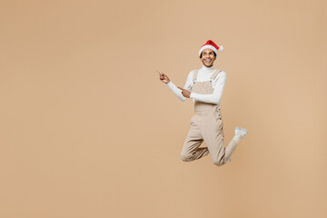 Fototapeta na wymiar Full body young african man in Santa Claus red Christmas hat jump high point index finger aside on workspace isolated on plain pastel beige background studio. Happy New Year 2022 celebration concept