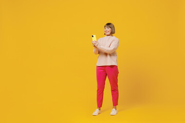 Full body elderly happy woman 50s wears pink casual knitted sweater using mobile cell phone chatting browsing internet isolated on plain yellow background studio portrait. People lifestyle concept