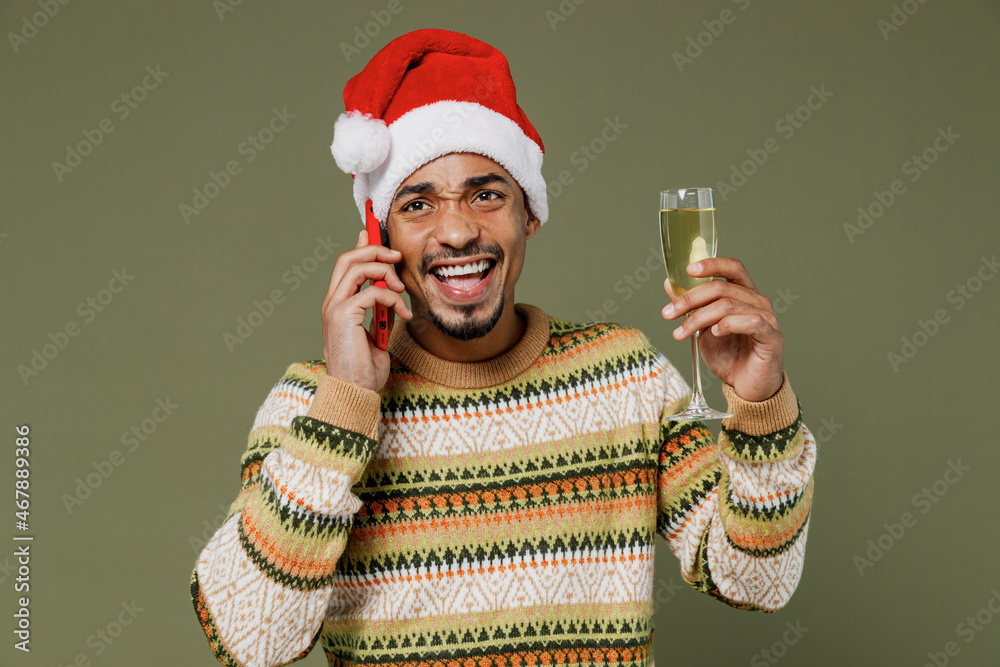 Wall mural Young african man in sweater red Christmas Santa Claus hat speak talk mobile cell phone hold champagne wineglass greet isolated on plain green khaki background Happy New Year 2022 celebration concept.