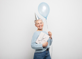 Amazed old woman holding blue balloon and gift box over white gray background.