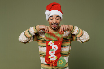 Young happy african man in knitted sweater red Christmas Santa Claus hat look at stocking gift sock isolated on plain green khaki background studio portrait. Happy New Year 2022 celebration concept.