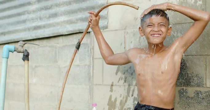 Slow motion scene of an Asian boy who is the farmer's son in a rural area uses a hose to pour water over his head to take a shower outside the house with a laugh and happiness.