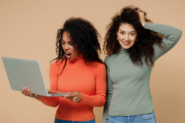 Two puzzled young curly black women friends 20s wear casual shirts clothes hold use work on laptop pc computer iterates over solution options isolated on plain pastel beige background studio portrait.