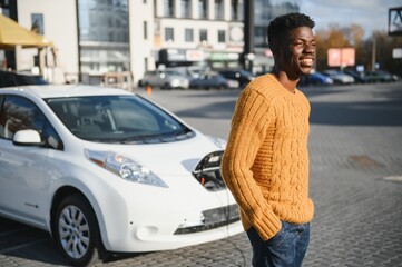 Fototapeta premium Urban, electric vehicle, eco concept. Young black skinned man, waiting for his electric car charging
