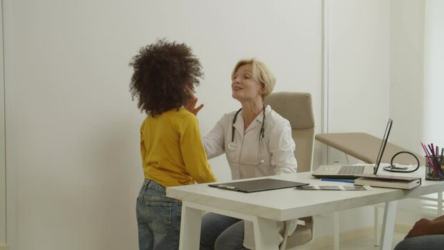 Caring attractive middle aged female general practitioner giving medical checkup, examining cute little multiracial boy sore throat while black family visiting doctor at hospital.