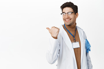 Smiling doctor in glasses and medical robe, pointing and looking behind at logo, healthcare advertisement, standing over white background