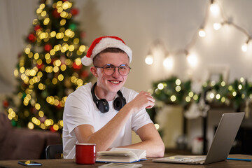 happy teenage student in a Santa hat and glasses uses a laptop for distance learning. a young man...