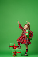 little girl years old with curly hair in golden deer horns, standing with gift boxes on green background in studio. child smiles happily and looks away and points finger. Advertising. copy space