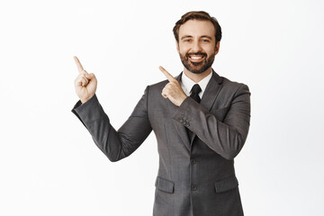 Portrait of successful businessman in suit, pointing fingers at upper left corner and smiling, showing advertisement, white background